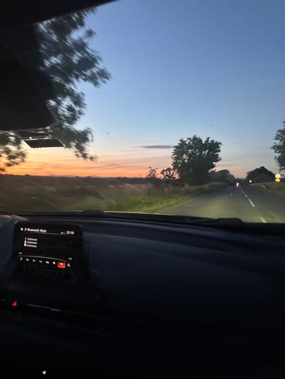 The image depicts a sunset viewed from inside a moving vehicle on a road near Elhovo, Bulgaria. The sky transitions from blue to orange hues, and the sun is setting over a landscape with trees and fields. The road ahead is illuminated by the vehicle's headlights, and a dividing line is visible. A dashboard screen shows music playing via Bluetooth. The interior of the vehicle is dark, with only portions of the dashboard and steering wheel visible.