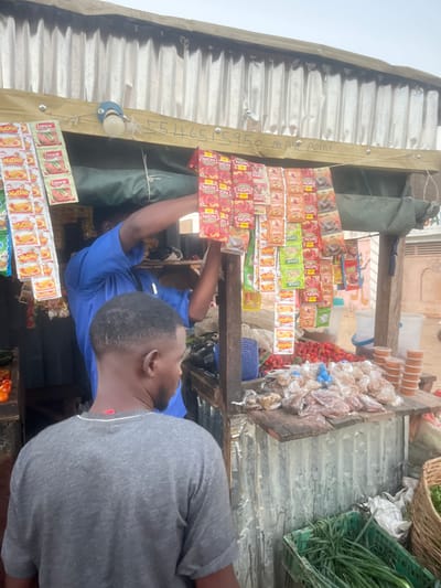 A person in a blue shirt is arranging sachets of tomato paste and instant noodles in a small shop. The shop appears to have a corrugated metal roof and wooden shelving. Other items for sale include packaged snacks, fruits, and vegetables. A second person wearing a grey shirt is partly visible in the foreground. There is a phone number visible on a wooden beam.