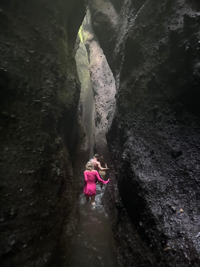 The image shows two people walking in a narrow canyon with dark, wet rock walls. The person in the foreground is wearing a bright pink dress, and the person behind them appears to be a shirtless man. Water is flowing around their legs. The canyon is dimly lit, with some light filtering in from above. This scene is likely from the Tukad Cepung Waterfall in Bangli, Indonesia, known for its unique cave-like waterfall and rock formations.