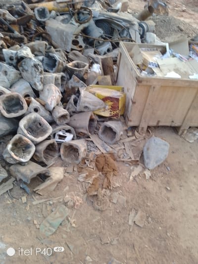 A disorganized heap of metal scraps and trash dominates the image. The background features remnants of machinery, including a steering wheel and other unrecognizable parts. A significant portion of the foreground is occupied by numerous hollow, conical metal objects with a rough, weathered texture. A wooden container filled with debris sits to the right. Debris scatters the dirt ground.