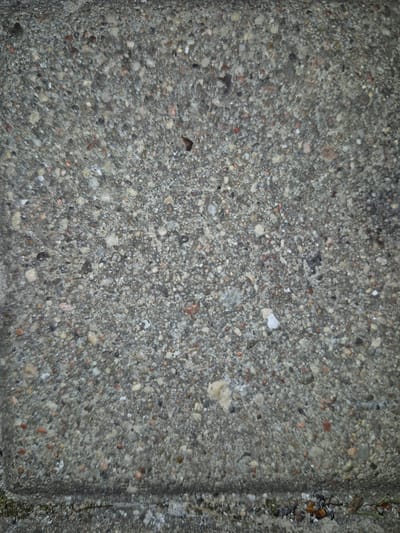 The image displays a close-up, overhead view of a grey concrete surface with embedded gravel. The texture is rough and appears to be a sidewalk or pathway. Small pebbles of various colors, including white, grey, red, and tan, are visible throughout the concrete matrix. In the lower portion of the image, a distinct horizontal line indicates the edge of a concrete slab, and a small amount of debris, including dry grass and possibly some dark-colored organic matter, is present along this edge. The overall lighting suggests an outdoor environment, possibly under overcast skies, with the concrete appearing slightly damp in places, reflecting small points of light. There are no people or animals present in the image.