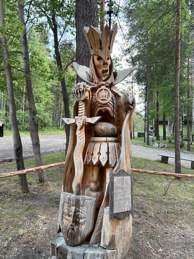 A wooden sculpture of a king or warrior figure stands in an outdoor setting, likely a park or historical site, at the Taltsy Museum, Tal'tsy, Russia. The figure is elaborately carved, featuring a crown, a skeletal face, a cape, and a sword. A shield with a carved face rests at its base. The sculpture is positioned in front of a line of trees, with a dirt path and grassy areas visible in the background. A wooden fence runs horizontally across the foreground, separating the viewer from the sculpture. The overall lighting suggests it is daytime, with overcast skies.
