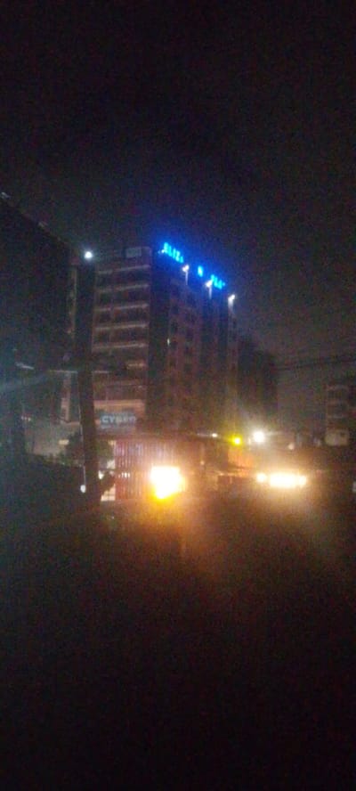 In Ndenderu ward, Kenya, at night, a multi-story building stands illuminated by bright blue neon lights spelling out "ELIZA N PLAZA" across its upper facade. The building, constructed of brick and concrete, has visible horizontal window lines or structural elements. Below the prominent signage, another sign is partially visible, appearing to read "CYBER". The street level is obscured by the darkness and the glare of vehicle headlights, with at least two vehicles visible with bright headlights. One vehicle in the foreground has its headlights illuminated, casting a strong yellow glow. Another vehicle's lights are visible further down the road. The scene suggests a commercial area with active businesses operating after dark, indicated by the illuminated signage and vehicle activity. The overall impression is of a typical urban nighttime scene in a commercial district.