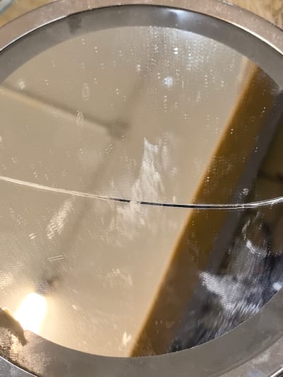 The image shows a close-up, angled view of a circular, mirrored surface, likely a tabletop or a large decorative mirror, showing numerous smudges, streaks, and dust particles. The mirror has a metallic rim. In the reflection, a bright light source, possibly a lamp, is visible on the left side, casting a glare. A blurred, warm-toned vertical element, possibly a wooden beam or furniture, is also reflected. A thin, dark, horizontal line, potentially a crack or a scratch, is visible across the mirrored surface. The background of the reflection is a muted, neutral tone. The scene is indoors, and given the general context provided, it is located in Novo-Talitsy, Russia.