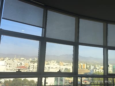 The image displays an elevated view of North Nicosia, Northern Cyprus, through a series of large, multi-pane windows with light-colored frames. Several light-grey roller blinds are partially drawn, covering the upper sections of most windowpanes to varying degrees.
Beyond the windows, an urban landscape unfolds under a clear, slightly hazy blue sky. The cityscape consists predominantly of multi-story buildings, primarily white, beige, or light grey in color. A distinct yellow/orange apartment building block is visible in the middle-ground to the right, and a building with green-tinted glass appears on the far right. In the distance, slightly left of center, a mosque with two minarets stands out. Scattered trees and green foliage are present among the buildings and in the lower foreground. A range of distant mountains, appearing blue-grey due to atmospheric haze, forms the horizon line. No individuals are visible. A digital timestamp "21/11/2025 09:09" is overlaid in the lower right corner of the image.