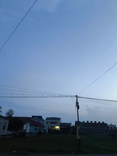 The image depicts a dusky sky over a semi-urban area in the city of Bireuenmeunasahblang, Indonesia. In the foreground, a grassy field with dark foliage stretches across the bottom of the frame. A utility pole with multiple wires crisscrossing the sky stands prominently to the right. Behind the field, a row of buildings is visible, including residential houses with pitched roofs and some larger, more modern structures. The lighting suggests it is either dawn or dusk, with the sky transitioning from a deep blue to a lighter hue. The overall impression is one of a quiet, developing locality as daylight fades.