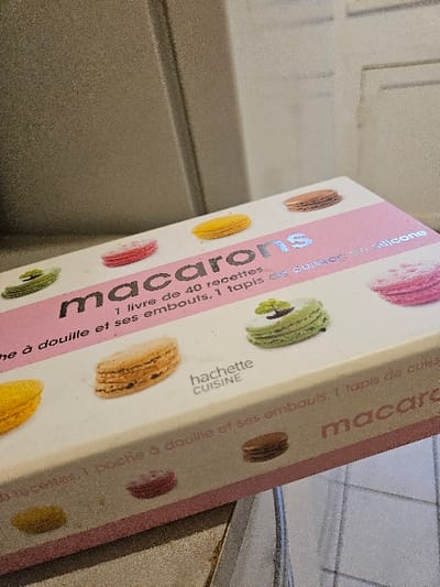 A rectangular white box, appearing slightly dusty, is displayed at an upward angle on a textured grey surface. The box features a prominent pink horizontal band across its upper face. Within this band, the word "macarons" is centrally printed in a reflective, possibly silver, typeface. Below the main title, smaller text in French reads "1 livre de 40 recettes" (1 book of 40 recipes) and "1 poche à douille et ses embouts, 1 tapis de cuisson en silicone" (1 piping bag and its tips, 1 silicone baking mat). Images of various colorful macarons are depicted on the box's surface, including pink, yellow, light brown, and green macarons, with the green one featuring a white decorative swirl. The "hachette CUISINE" logo is visible on the box. The background consists of a light grey, vertically textured surface, likely an appliance door or wall. No individuals are present, and no actions or interactions are occurring. The scene takes place indoors.