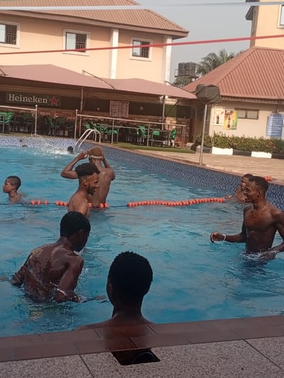 Several young men are in a swimming pool, some waist-deep, others closer to the edge. The pool is lined with an orange buoy line. In the background, there's a building with a "Heineken" sign, tables, and chairs, suggesting a bar or restaurant. Further back, there are two-story buildings with windows and a water tank. The scene is likely set at a resort or recreation area, capturing a moment of leisure.