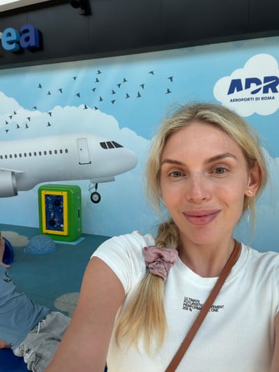 This is a close-up, vertically oriented selfie of a young woman with blonde hair pulled into a braid. She is wearing a white ribbed t-shirt and a brown strap is visible across her shoulder. She has light skin, green eyes, and a slight smile. A small, star-shaped earring is visible in her left ear. The background is a mural depicting a blue sky with white clouds and numerous silhouetted birds in flight. A large white airplane is painted on the mural, with its front section and a landing gear wheel visible. To the right of the airplane, there is a green interactive panel with a yellow frame and a blue patterned interior, and a blue mat is on the floor. In the lower left corner of the image, large blue and light blue letters, possibly part of a sign, are visible. The text "APR AEROPORTI DI ROMA" is visible on a white cloud shape in the upper left portion of the mural. The text "ULTIMATE FUTURISTIC PERFORMANCE 200 %" is printed on the woman's t-shirt. The setting appears to be an indoor area, possibly a children's play area or waiting area, at an airport in Rome, Italy. The lighting suggests it is daytime.