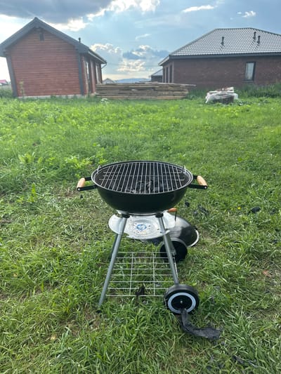 In Мокино, Russia, a black charcoal grill with a silver stand sits on a grassy lawn. The grill is open, revealing charcoal inside. The stand has two wheels, one of which has a black plastic bag partially covering it. In the background, there are two buildings, one made of red wood and the other of brick, and a pile of lumber. The sky is cloudy.