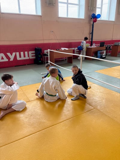 This image captures an indoor sports facility, likely a dojo or gymnasium, in Kuyeda, Russia, showing a break or administrative area during a youth sports event. The foreground features a yellow tatami mat floor with a section of grey-green matting visible further back, separated by a white boundary line and a low white railing.
Five children are present in the scene. In the left foreground, a boy with light brown hair, wearing a white judogi with a green belt, sits cross-legged, facing right, with his left hand near his chin. Adjacent to him, a second boy, also in a white judogi with a green belt, sits on the mat, facing left with legs extended. To the right, a third boy with dark hair, wearing a black hooded sweatshirt and white pants with grey socks, is in a low crouch, looking towards the camera with an open mouth. Partially obscured behind the second boy, a fourth boy in a blue and green tracksuit is kneeling on the mat, facing away. In the background, near the administrative tables, a fifth boy with dark curly hair, wearing a white judogi, stands holding a blue balloon.
The background wall is painted a light peach color. A red banner with white lettering and stylized athletic figures runs along the lower portion of the wall. Partially visible text on the banner includes "ДУШЕ" and "УК". High on the wall are large, multi-pane windows covered by a fine white mesh or netting.
An administrative area is set up with two light-colored wooden tables. On the right table, a black laptop and water bottles are visible. A digital scoreboard, predominantly black with blue and white elements and displaying "W" and "W", rests on the floor near this table. Between the tables, a blue vertical pole is topped with three balloons: two blue and one red. On the left table, small dark objects resembling microphones are present, and a tall black speaker with prominent circular blue, green, and red LED lights is positioned on the floor nearby. The children on the mat appear to be resting or observing, while the boy in the black hoodie looks directly towards the viewer.