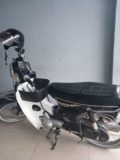 A black and white Honda motorcycle is parked indoors, likely in a garage or covered area, in Geudonggeudong, Indonesia. The motorcycle is upright, supported by its kickstand, and appears to be in good condition. A black helmet with a clear visor is placed on the handlebars. The motorcycle features a black seat with white stitching and a white front fairing. The word "HONDA" is visible on the side of the motorcycle. The background consists of a plain gray wall and a tiled floor. The lighting is even, suggesting an indoor, possibly shaded, environment. No people are present in the image.