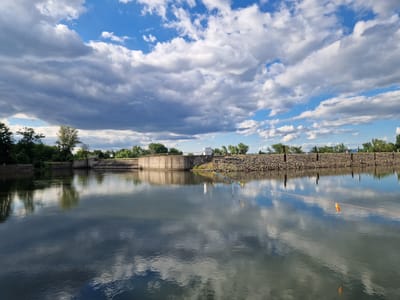 The image shows a tranquil river scene under a dramatic sky filled with both dark and white clouds, casting reflections on the water's surface. In the background, a concrete structure, likely a dam or floodgate, spans the river. The embankment of the dam is lined with a rocky wall and some vegetation. A string of colorful small flags runs along the edge of the water, suggesting a festive or demarcated area. There are no people prominently visible in the image, contributing to the serene atmosphere. The location is identified as Pancharevo, Bulgaria.