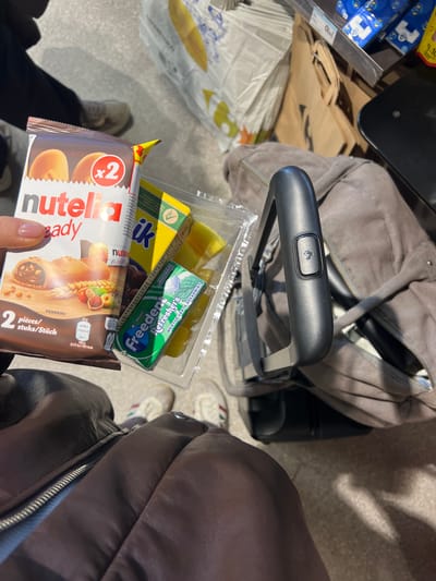The image appears to be a close-up, first-person perspective shot of a collection of items held in someone's hand or positioned on a surface next to a suitcase.

The main items visible are:
- A package of "Nutella B-ready" pastries (x2).
- A package of "Melk" cereal bars.
- A pack of "Freedent Refreshers" chewing gum.
- A small container with what appears to be pineapple chunks.

The items are being held next to a grey suitcase with a black handle. Other items such as paper bags are also visible on the side, and a person wearing a brown jacket can also be seen.

The image most likely took place in the city of Paray-Vieille-Poste, France.