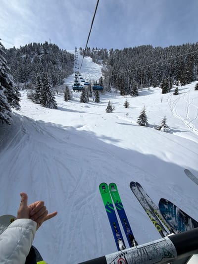 This image presents a first-person view from a ski lift ascending a snow-covered mountain in Les Allues, France. In the immediate foreground, the black safety bar of the ski lift is visible, displaying a worn white and grey patterned sticker. Suspended from the bar are two pairs of skis. The pair on the left features a distinct green and blue design, branded "DYNASTAR." The pair on the right exhibits a mixed pattern, with one ski showing a black and yellow design and the other a blue and white abstract pattern. To the left of the frame, a person's left arm, clad in a white ski jacket sleeve, extends forward, with the hand forming a "shaka" gesture (thumb and pinky extended).
The environment below consists of a wide, groomed ski slope covered in deep snow, showing numerous parallel ski tracks. Dense evergreen forests, with trees heavily laden with snow, flank the ski slope on both sides and extend up the mountain. In the middle ground, a series of blue-grey chairlift chairs are visible ascending the mountain along a steel cable. The sky above is largely clear and blue, with scattered white clouds. Long shadows stretch across the snow, suggesting specific sun angle.