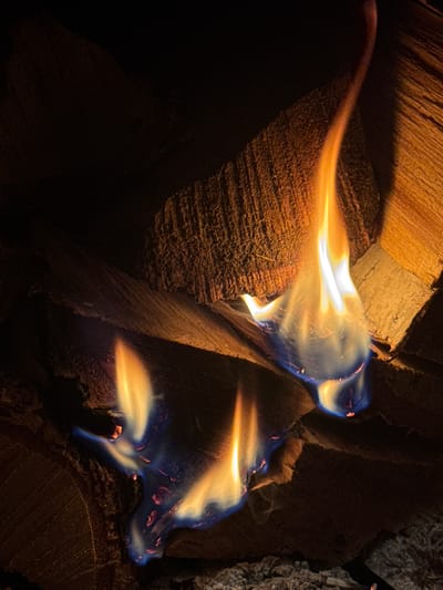The image depicts a close-up view of a bonfire with flames engulfing pieces of wood. The fire displays vibrant colors ranging from bright yellow and orange to a deep blue at its base, indicating intense heat. The wood logs are textured and appear to be dry, stacked against each other, forming the structure of the fire. The background is dark, suggesting the photo was taken at night or in a dimly lit environment. There are no people visible in the image. The scene is of a natural phenomenon, likely a controlled fire for warmth or ambiance. The location in Городецкий ауылдық округі, Kazakhstan, implies this could be a campfire in a rural or semi-rural setting.