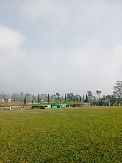 The image shows a grassy soccer field in Kudawa, Sri Lanka under a cloudy sky. In the background, behind a stone wall, are several trees and buildings, some partially obscured by mist. Along the side of the field is a small, green covered seating area with red seats. A soccer goal is visible on the right side of the frame. The field itself has visible white lines, indicating markings for the game.
