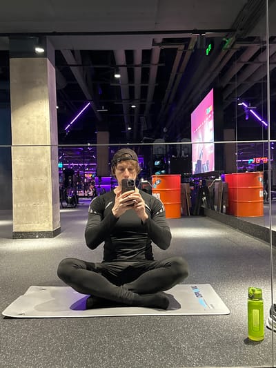 The image depicts a reflection of a person taking a photo of themselves in a mirrored room. The person is wearing a black long-sleeved top and black leggings, sitting cross-legged on a white yoga mat. A green water bottle sits to the side.

The background is the reflection of a gym-like space with weights, orange barrels, and a large screen displaying an image. The architecture suggests an industrial or warehouse-style building, featuring visible pipes, metal frameworks, and concrete columns. The lighting includes purple neon strips and individual spotlights.

The overall setting is modern and fitness-oriented. There is no definitive visual information to identify the exact location in Murino, Russia.