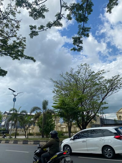 The media file shows a street scene, likely in Mandar Sari, Indonesia, on a bright, cloudy day. In the foreground, a white car is parked on the right side of the road. Next to it, a person wearing a dark helmet and jacket is riding a motorcycle, visible from the side. The road has a distinct black and white striped curb and yellow lane markings.
Behind the parked car and motorcycle, a row of palm trees and other green foliage line the street. Further back, there is a building with ornate architectural details, possibly a mosque or a historic structure, painted in a light cream color with blue accents on the windows.
The upper portion of the image is dominated by the sky, filled with a mix of bright blue patches and white, fluffy clouds, suggesting a day with fluctuating sunlight. Several branches of trees with green leaves extend into the frame from the left, partially obscuring the sky and some of the buildings. A street light is visible in the lower part of the image, near the center. The overall atmosphere is that of a typical urban or suburban street scene in a tropical or subtropical region.