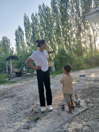 In Тайгараев, Kyrgyzstan, a woman in a white t-shirt and black pants stands with her hands on her hips, looking to the right. A young child in a tan shirt and patterned shorts faces away from the camera, towards the woman. The environment is rural, with tall trees in the background and a dirt and concrete ground. There is a small, open-sided structure with a dark roof to the left of the woman, and a metallic object resembling a watering can or a samovar stands near the child. Sunlight filters through the trees, creating a warm, late afternoon atmosphere.