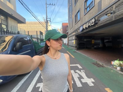 A young woman wearing a grey tank top, a green baseball cap, and pearl jewelry is taking a selfie on a street in Daegu, South Korea. The street has green and white markings, suggesting a bike lane or pedestrian walkway. A blue pickup truck is parked on the left side of the image, and a building with Korean signage is visible on the right. The building appears to be a commercial establishment, possibly a business or apartment complex, with an entrance to a parking garage. The overall atmosphere is bright, with clear skies indicating daytime. There are trash bags piled up near the building entrance.