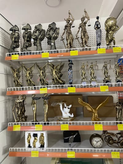 This is a medium shot of a retail display of various decorative statues and figurines, likely in a shop. The items are arranged on white wire shelves, with orange vertical supports visible. The overall color palette is dominated by gold and bronze tones from the statues, interspersed with the white of the shelves and the orange of the supports.
The subjects are primarily inanimate objects. On the left, there are several golden statues of female figures holding scales and swords, commonly representing Lady Justice. Below these are more abstract, dark-colored figures with hands covering their faces. Further down, there are more golden, winged figures.
In the center, a large golden eagle statue is prominently displayed. To its right, a white dog figurine wearing sunglasses and a bowtie stands out. Further to the right, a vintage-looking clock with Roman numerals is visible. Other items include metallic ducks and abstract golden forms.
The scene appears to be an indoor retail environment, possibly a home decor or gift shop. There are price tags visible on some of the items, with text that includes "GOLDEN," "EAGLE STATUE," "BULLDOG TR," and numerical values like "17.300 N," "28,700 N," and "900N." The location context provided states "Galadimawa Village, Nigeria," suggesting this is a shop in that region. There is no discernible activity or event taking place, and the image is static. The lighting is neutral and appears to be from overhead fixtures, indicating it could be daytime or evening.
Notable details include the metallic finishes of the statues, the variety of figures from classical representations to more modern and whimsical designs, and the presence of price tags indicating commercial activity.