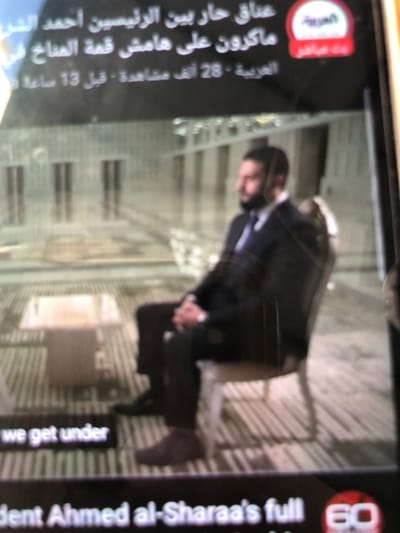 The image displays a photograph of a screen showing a news broadcast, presumably depicting an interview or statement from a formal indoor setting identified as being in the city of Kafr Sousa Municipality, Syria. In the foreground, a man with dark hair and a trimmed dark beard is seated in profile, facing left. He is dressed in a dark suit jacket over a light shirt with a dark, possibly purple, tie. His hands are clasped, resting between his knees, and he wears dark trousers and dark brown low-top shoes. He sits on a chair with a light-colored frame and a dark upholstered back and seat. To his left, a low, square table with a light-colored surface and base is positioned on a patterned carpet featuring vertical stripes and geometric designs. The background reveals a grand interior space with high ceilings and what appear to be multiple tall, narrow architectural features, possibly columns or windows, suggesting a large hall or formal room. Overlays on the broadcast screen include Arabic text and the Al Arabiya channel logo in the upper right, and partial English text at the bottom, including "we get under," "sident Ahmed al-Sharaa's full," and the "60 Minutes" logo in the lower right corner.