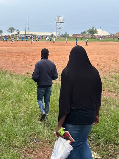 In Du, Nigeria, on an overcast day, two individuals walk on a grassy area adjacent to a dirt soccer field where a game is in progress. The person in the foreground, wearing a black headscarf and dark top, is holding a white plastic bag and a red object. The person in the middle ground, a male, wears a dark blue jacket and jeans, and walks away from the camera towards the soccer field. Numerous players in various colored uniforms are actively engaged in a soccer match on the red dirt field. In the background, buildings, a water tower, and a tall communication tower are visible under a cloudy sky.