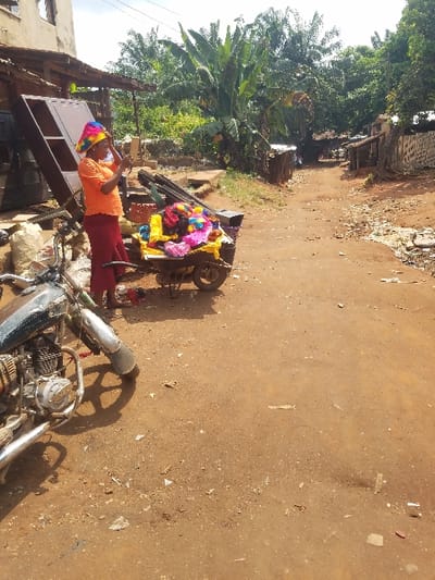 The scene is an unpaved reddish-brown dirt road in the city of Nnobi, Nigeria, captured during daylight. In the left foreground, a dark, weathered motorcycle is parked on the dirt. Adjacent to it, an adult female stands barefoot, wearing an orange short-sleeved top, maroon trousers, and a brightly colored, multi-patterned conical hat. She holds a small rectangular mirror in her hands, looking into it. Beside her is a two-wheeled handcart loaded with numerous brightly colored fabric items, predominantly yellow, pink, purple, and blue. Behind the woman and cart, an incomplete concrete block building rises, with dense tropical foliage, including large banana plants, visible beyond. The dirt road extends into the background, flanked by various simple structures with corrugated metal roofs and additional green vegetation, before curving slightly to the right and receding into the distance. Scattered debris is present on the road surface.