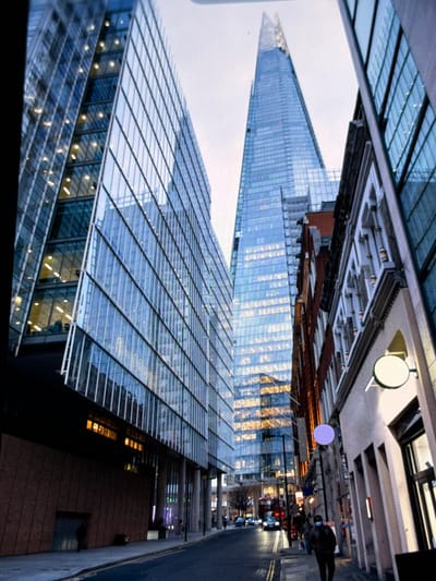 This image depicts a street scene in London, United Kingdom, not Maidstone, prominently featuring The Shard skyscraper.
In the background, The Shard, a tall, glass-clad pyramidal skyscraper, stands centrally against an overcast, light grey sky. On the left, a multi-story modern building with a blue glass curtain wall displays numerous illuminated interior office spaces. Its lower section features a dark, textured facade. On the right, a sequence of buildings includes a structure with a light-colored, decoratively carved upper facade and a plain lower section, from which a circular white sign projects. Further along this street, a building with a red brick facade is visible, above which a circular purple light is mounted.
The foreground shows an asphalt street with visible double yellow lines, flanked by concrete pavements. On the right-side pavement, a pedestrian wearing a dark coat and a blue face mask walks towards the viewer. Additional pedestrians are discernible further down the street. Vehicular traffic, including a red double-decker bus and several cars with illuminated headlights, is present in the middle ground. Multiple streetlights are active. The ambient lighting from the sky is subdued, consistent with an overcast day or early evening.