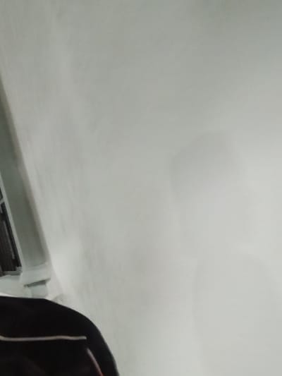 The image shows a close-up of a white wall, with a dark object in the lower left corner, possibly a piece of clothing. The wall appears to be textured and is lit from the left, casting a subtle shadow. The overall impression is one of an indoor environment, possibly a room, with a focus on the wall's surface. There is no indication of the city of Tacarigua, Venezuela, or any other specific location.