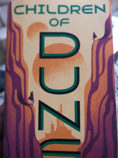 The image displays the front cover of the book 'Children of Dune' by Frank Herbert, held by a human hand. The cover features a stylized illustration on a pale yellow-beige background. Two orange celestial bodies are visible in the upper center. The title 'DUNE' is prominently displayed vertically in large, black letters outlined in dark teal, with 'CHILDREN OF' in smaller green text positioned above. The illustration includes undulating, multi-toned orange and purple rock formations or sand dunes that frame the central text. Two small, cloaked humanoid figures are depicted on these formations: one on the lower-left, facing right and casting a long shadow, and another on the upper-right, facing left. An orange silhouette of a city, featuring spires and domes, is integrated into the lower section of the letter 'N'. A human hand, with dark skin, grips the lower-left side of the book. The background behind the book is blurred, indicating an indoor setting. This image was captured in Muguga ward, Kenya.