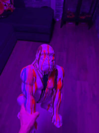 A painted, stylized human figure lies on its side on a wooden floor. It’s patterned with dripping paint in red, black, and blue. A hand holds it. The scene is illuminated with a purple light. A dark gray couch is in the background. A bookshelf is also visible, although it's out of focus due to the low light.