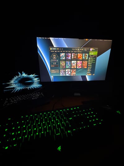 The image depicts a dimly lit desk area with a computer screen and a Razer brand gaming keyboard with green backlighting. On the screen, a user interface is visible, displaying a profile page within the "League of Legends" client. The profile contains various icons, champion skins, and player stats. The desktop background features a modern, abstract design. Based on the UI language observed in the screenshots the computer is operating in french. The UI contains the title "Musique Fichier Edition Morceau Présentation Commandes Compte Fenêtre Aide" which indicates the UI is running on a MacOS system.
There is not any information within the image to ascertain the location of the image to be Les Ulis, France.