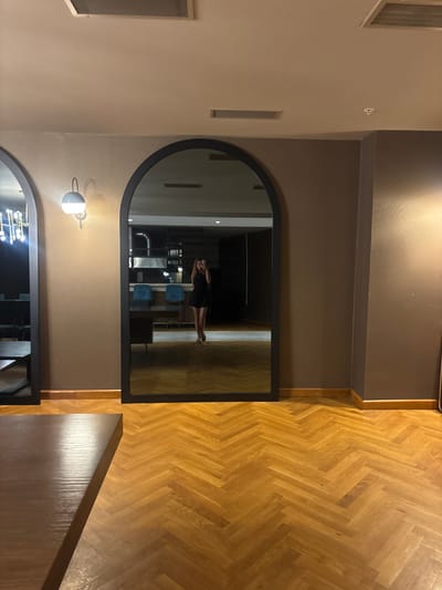 An arched mirror reflects a woman taking a photo with her phone. The woman is wearing a black dress and standing in a room with dark grey walls and a wooden herringbone floor. There are blue chairs and a dark table visible in the reflection, suggesting the location may be a restaurant or lounge. The room also features a light fixture on the wall.