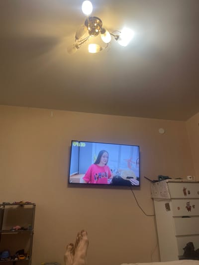 The image shows an indoor scene, likely a living room, with a television mounted on the wall. The TV displays a program with a young girl wearing a pink t-shirt. The room is dimly lit, with a ceiling fan with multiple lights visible at the top of the image. To the left of the television, a grey shelving unit holds various items, including toy cars. To the right of the television, a white dresser with drawers is decorated with Christmas-themed stickers. In the foreground, a pair of feet are visible, suggesting someone is sitting or lying down, watching the television. The walls are painted a neutral color. The location is identified as Laishevo, Russia.