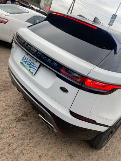 The image captures the rear view of a white Range Rover, likely a Range Rover Evoque, parked on what appears to be a street or unpaved lot in Agege, Lagos, Nigeria. The vehicle features sleek black accents, including a black roof and rear bumper, contrasting with its white body. Its distinctive LED taillights are illuminated bright red, and the "RANGE ROVER" lettering is visible across the tailgate, along with a "LAND ROVER" emblem on the right side.

A Nigerian license plate, clearly displaying "LAGOS," "CENTRE OF EXCELLENCE," and the alphanumeric code "AAA-840HU," is mounted at the center of the rear. To the right of the car, a black tire leans against its bumper. Another white car, a sedan, is partially visible behind the Range Rover, suggesting a crowded parking or street environment.

The ground beneath the vehicles is a rough mixture of dirt and gravel. In the background, against an overcast sky, overhead wires crisscross, and a banner or billboard is visible with partial text including "Hope" and "DOOR TO DOOR." The overall lighting suggests a cloudy day, with no harsh shadows.