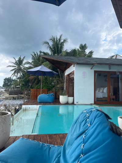 This image depicts a serene outdoor scene at a resort in Nusa Penida, Indonesia. In the foreground, a bright blue beanbag chair is partially visible, facing a swimming pool. The pool itself is a vibrant turquoise color, with a portion of it in the foreground appearing shallower and lighter, possibly due to sunlight or the pool's design. The pool is bordered by a wooden deck, upon which rests another blue beanbag chair and a large blue umbrella providing shade. Beyond the pool and deck, a white building with wooden-framed glass doors and windows is visible. The interior of the building reveals a bedroom setting with a neatly made bed adorned with pillows. Palm trees and lush green vegetation are scattered throughout the background, hinting at a tropical setting. In the distance, a glimpse of the ocean and rocky coastline can be seen, under a cloudy sky. The overall atmosphere is one of relaxation and tropical vacation.