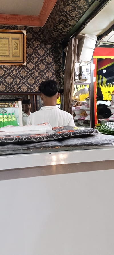 The scene takes place inside a shop in Kecamatan Kutawaringin, Indonesia, located at coordinates -6.988 latitude and 107.533 longitude. A man with short black hair, wearing a white shirt, is standing with his back to the camera facing a counter. He appears to be waiting to be served. In front of him is a white counter with a stack of black plastic bags on top.  A man wearing a white shirt is partially visible in the background.  A large framed picture hangs on the wall to the left of the counter.  A refrigerator is visible on the left, with bottles of green liquid inside.  In the background, a rack of goods can be seen on the right.  The walls are decorated with floral wallpaper. There is an open doorway on the right side.  It is unclear what the business is selling, but it is likely a food or drink establishment.