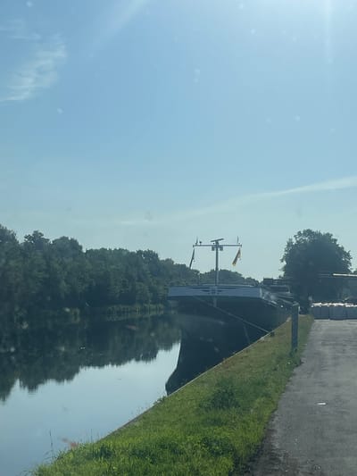 The image shows a canal with a barge docked along the right bank. The canal is lined with lush green trees on the left bank, their reflections clearly visible in the calm water. The barge, a long, dark vessel, is positioned parallel to the bank. A tall mast with two flags and some equipment is mounted on the barge. To the right of the barge, there is a grassy embankment leading to a paved area where stacks of what appear to be pallets of goods are stored. The sky is a bright, clear blue with some faint wispy clouds and lens flare from the sun. The location appears to be in Edewecht, Germany, given the context provided.