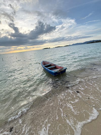 In Baan Plai Laem, Thailand, a small, blue and red rowboat rests partially beached on a sandy shore. The boat, unoccupied, is tethered by a rope to an unseen point, with its stern facing the water. Gentle waves lap at the sand, creating a foamy shoreline. The sea stretches out to the horizon, where a hazy line of distant islands and mountains can be seen. The sky above is a mix of dramatic clouds, with patches of sunlight breaking through, suggesting either dawn or dusk. The water is calm and reflects the muted light of the sky. There are no people visible in the image.