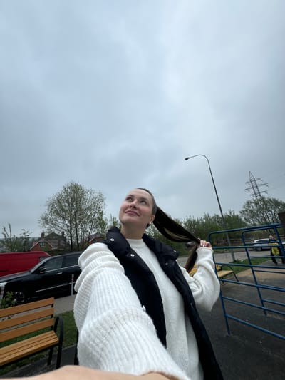 A young woman wearing a white sweater and black vest stands outdoors on an overcast day. She is holding her long, dark hair in her hands. In the background, there is a street with a street lamp and power lines overhead. There's also a blue metal fence, trees with sparse leaves, parked cars, and wooden benches. The location is identifiable as Akulovo in the Gp Kubinka region of Russia based on the architectural style of the buildings.