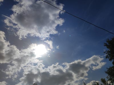 The image depicts a bright, sunny sky with scattered cumulus clouds. The sun is prominently visible on the left side of the frame, partially obscured by clouds, casting rays across the sky. A dark, thin wire stretches diagonally across the upper portion of the image. The lower right and bottom edges of the frame show the tops of dark green trees and foliage, suggesting an outdoor setting. The overall scene is a natural landscape, likely a clear day with dynamic cloud formations. The image does not contain any people, identifiable buildings, or urban elements that would pinpoint a specific location within the city of Tosno, Russia.