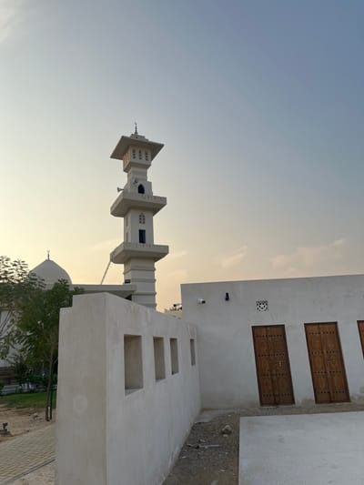 The image presents an outdoor architectural scene located in Sharjah, United Arab Emirates, under a clear sky exhibiting a gradient from a pale yellow-orange near the horizon to a light blue overhead, indicative of either sunrise or sunset. A multi-tiered, light-beige minaret with a square base stands prominently in the left midground, featuring recessed decorative elements, multiple window openings, and a crescent moon finial at its apex. Two dark loudspeakers are affixed to a lower tier of the minaret. To the left of the minaret, partially obscured by a green-leafed tree, is a light-colored mosque dome. In the foreground, a light-beige stucco-finished wall with four regularly spaced, rectangular openings extends from the left toward the center. To the right of this wall, a light-beige, single-story building is visible, featuring at least three tall, ornately carved dark wooden doors with metal hardware. A decorative square vent with a star motif is positioned above one door. A white dome security camera is mounted on the upper left portion of this building, with a dark conduit running vertically beneath it. The ground in the immediate foreground comprises a concrete slab, transitioning to unpaved earth with scattered debris between the wall and the building, and a paved pathway bordered by green grass and a small tree to the far left. No individuals or active interactions are present in the scene.
