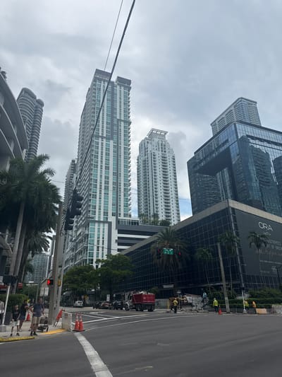 The image captures a street view in Miami, United States of America. The scene features several modern high-rise buildings with glass facades, typical of an urban setting. In the foreground, a street is visible with traffic lights, road markings, and construction cones, suggesting ongoing work or road maintenance. A few people are present on the sidewalk, some appearing to be construction workers in high-visibility vests, while others are casually dressed pedestrians. Palm trees are interspersed among the buildings, adding a tropical element to the urban landscape.
The sky is overcast, indicating a cloudy or potentially rainy day, and the lighting is diffused. The image is oriented vertically, with the buildings appearing to stretch upwards. Visible text on a large billboard on one of the buildings includes "ORA" and "RESIDENCES," along with an address and contact information, hinting that this might be a development or sales office. The overall atmosphere is that of a busy urban environment, potentially during daytime, with some activity related to construction or urban development.