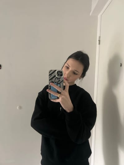 An eye-level mirror selfie captured indoors in the city of Cachan, France, depicts a female individual. The subject, a young adult with dark hair pulled back, is wearing a black crew-neck sweatshirt and a silver cross necklace. Her left hand, adorned with a gold-colored ring on the ring finger and a darker band on the middle finger, holds a smartphone. The smartphone, an iPhone model with a triple-lens camera system, is encased in a clear cover featuring a black and grey wavy pattern, with a blue card visible behind the phone within the case. The background, visible in the mirror reflection, consists of plain white walls. To the left of the subject in the reflection, a red rectangular object and a white circular object are mounted on the wall. To the right of the subject in the reflection, a white door with a visible metallic hinge and a dark circular fitting is present.