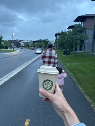 A first-person perspective shot shows a hand holding a disposable coffee cup as a man and child walk away on a paved pathway. The man wears a red and white plaid shirt and a green cap. The child, seen from the back, wears a pink jacket. The pathway runs alongside a road with a few cars and buildings in the background. The sky is overcast. The scene is likely in Gatineau, Canada, given the context. The coffee cup has a green circular logo with bamboo imagery and text that reads "10 BAMBOO BAMBOO BAMBOO BAMBOO NATUREL ET NON BLANCHI."