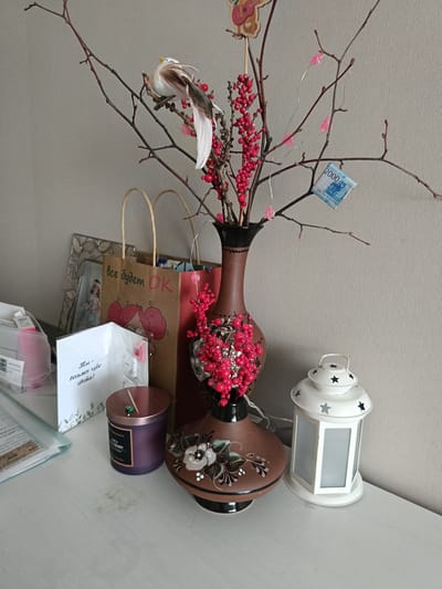 The image shows a still life composition on a white surface, possibly a table or shelf. The scene includes a brown vase adorned with red berries and artificial branches, featuring a decorative bird and a 2000 Russian Ruble bill attached to one of the branches. Alongside the vase, there's a white lantern with star-shaped cutouts, a purple candle, a framed photograph, a shopping bag with Russian text, and a greeting card also in Russian. The environment appears to be indoors, with a neutral-colored wall in the background. The text on the bag reads "Все будет ОК" (Everything will be OK). The card says "Ты - восьмое чудо света!" (You are the eighth wonder of the world!). The candle has the word "SPICY" and "BLACKBERRY" written on it. The bill is clearly labeled "ДВЕ ТЫСЯЧИ РУБЛЕ" (Two Thousand Rubles). There is no information to infer that the image was taken in Serpukhov, Russia.