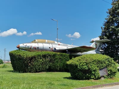 The image shows a retired jet aircraft displayed on a lawn in Svilengrad, Bulgaria. The aircraft is silver with red markings and appears weathered. It is positioned behind two trimmed hedges. A metal plaque is visible on the front right hedge. In the background, a tall tree, power lines, and buildings are visible under a clear blue sky with scattered white clouds. The scene is illuminated by bright daylight, suggesting it was taken during the day. No people are present in the frame.