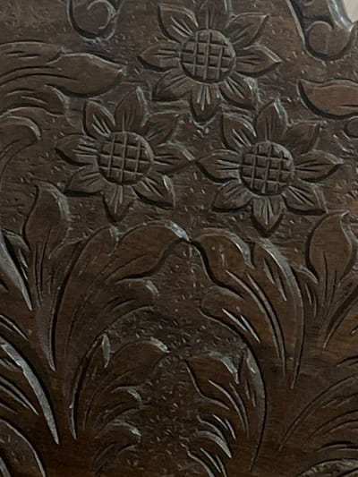 The image shows a close-up of a dark wood carving featuring floral and leaf motifs. Two stylized sunflower-like flowers are prominently displayed, each with a grid pattern in the center and carved petals radiating outwards. Flowing leaves and scrollwork fill the spaces between the flowers, creating an ornate decorative pattern. The carving appears to be old, with the wood exhibiting a rich, dark patina. The texture of the wood grain is visible beneath the carved details, suggesting a natural material. The lighting highlights the depth and dimension of the carving. This carving is likely part of a piece of furniture, architectural element, or decorative object found in Villa de Cura, Venezuela.